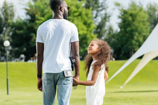 Vista trasera del policía afroamericano y su hija tomados de la mano en el parque de atracciones - foto de stock