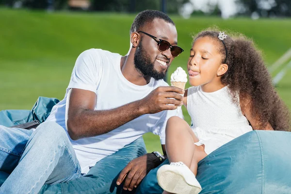 Porträt eines afrikanisch-amerikanischen Vaters, der seiner Tochter Eis auf Sitzsäcken im Park reicht — Stockfoto