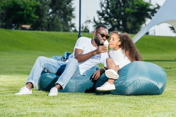 African american father giving ice cream to daughter on beanbag chairs in park — Stock Photo