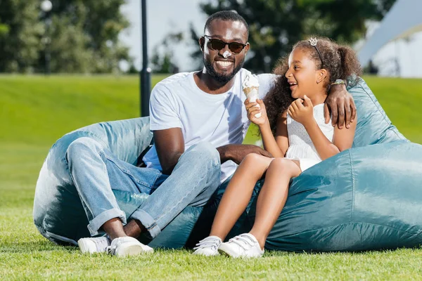 Sonriente afroamericano padre e hija con helado divertirse en frijoles sillas en el parque - foto de stock
