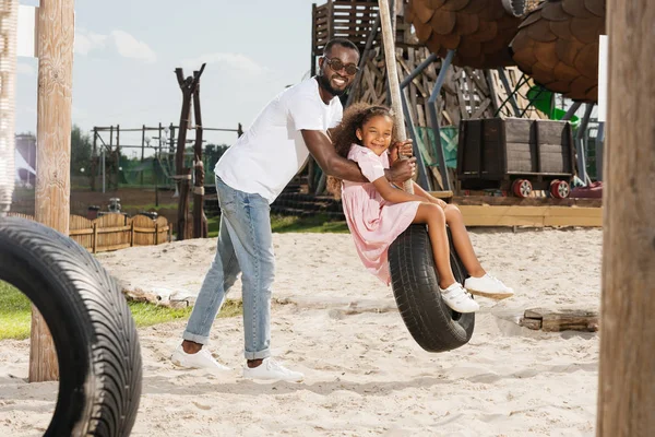Africano americano padre y hija en neumático swing en parque de atracciones - foto de stock