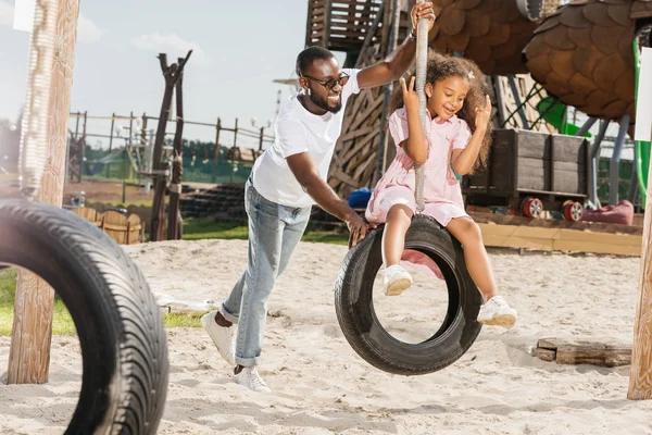 African american father and daughter on tire swing having fun on playground — Stock Photo