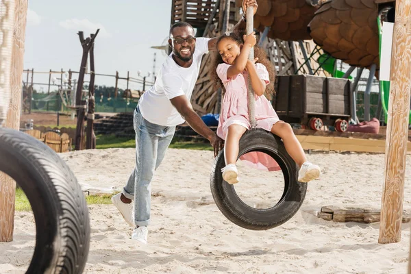 Щасливий афроамериканський батько штовхає дочку на гойдалки на шинах в парку розваг — стокове фото