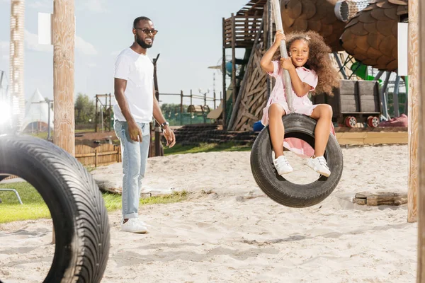 Afro-americano pai de pé perto sorrindo filha no pneu swing no parque de diversões — Fotografia de Stock