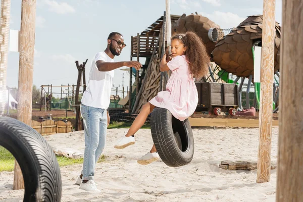 African american father catching daughter on tire swing at amusement park — Stock Photo
