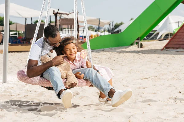 Afro-americano pai tocando filha bochecha no teia aranha ninho balanço no parque de diversões — Fotografia de Stock