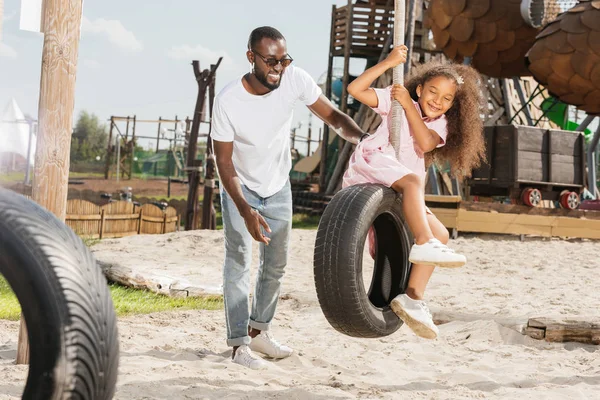 Smiling african american father standing near daughter on tire swing at amusement park — Stock Photo