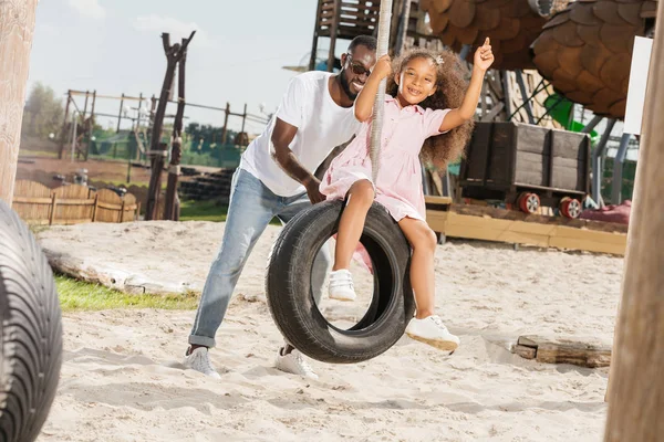 Africano americano padre empujando hija en neumático swing en parque de atracciones - foto de stock