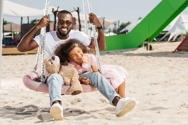 Lächelnder afrikanisch-amerikanischer Vater und Tochter auf Spinnennetz-Nestschaukel im Freizeitpark — Stockfoto