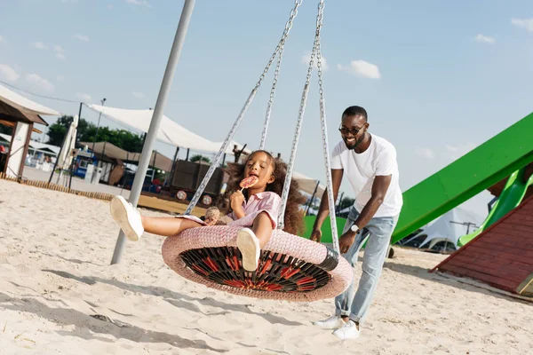 Happy african american daughter with lollipop on spider web nest swing at amusement park — Stock Photo