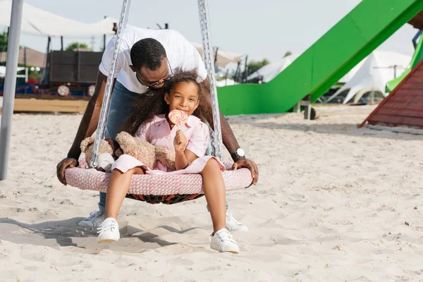 African american father pushing happy daughter with lollipop on spider web nest swing at amusement park — Stock Photo