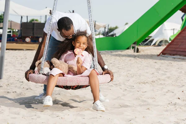 Afrikanisch-amerikanischer Vater schubst glückliche Tochter mit Lutscher auf Spinnennetz Nestschaukel im Freizeitpark — Stockfoto