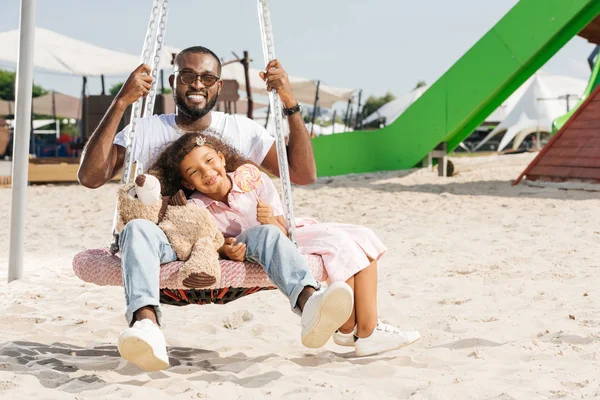 Feliz pai e filha afro-americanos no balanço do ninho de teia de aranha no parque de diversões — Fotografia de Stock