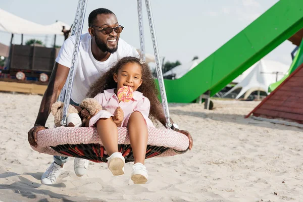 African american father pushing daughter on spider web nest swing with lollipop at amusement park — Stock Photo