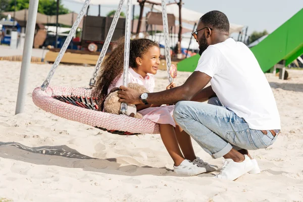 Africano americano padre sentadilla cerca hija en araña web nido swing en parque de atracciones - foto de stock