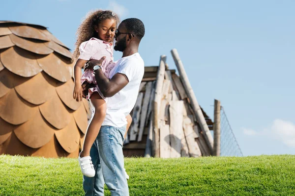 Africano americano padre holding adorable hija en verde colina en parque de atracciones - foto de stock