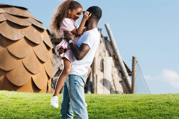 Père afro-américain tenant fille heureuse sur la colline verte au parc d'attractions — Photo de stock