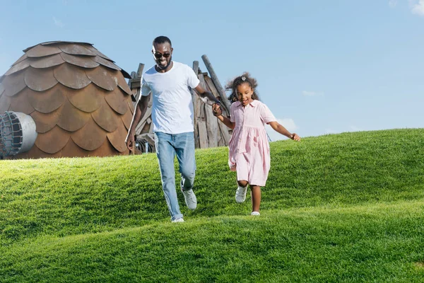 Afro-americanos padre e hija tomados de la mano y corriendo en la colina en el parque de atracciones - foto de stock