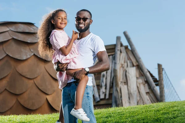 Smiling african american father holding daughter on green hill at amusement park — Stock Photo