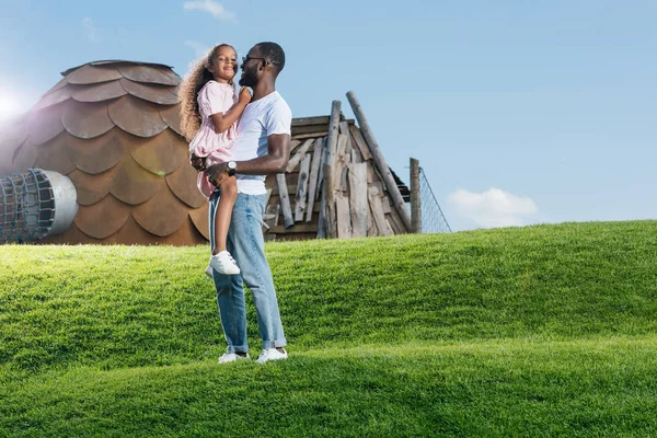 Alegre afroamericano padre holding hija en verde colina en parque de atracciones - foto de stock