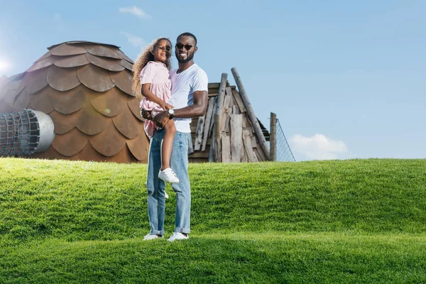 Африканский американский отец держит дочь на зеленом холме в парке аттракционов и смотрит в камеру — стоковое фото