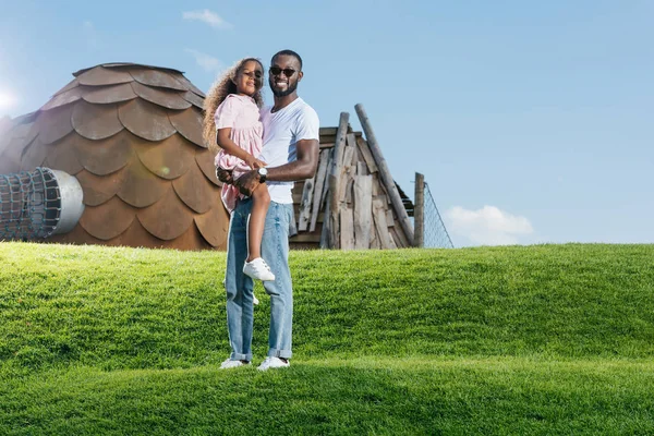 Africano americano padre holding hija en verde colina en parque de atracciones y mirando cámara - foto de stock