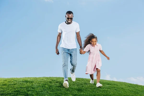 Afro-americanos padre e hija tomados de la mano y caminando en verde colina - foto de stock