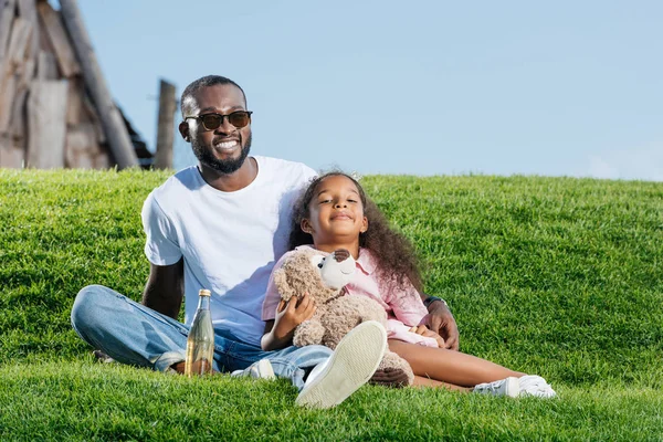 Heureux père afro-américain et sa fille assis sur la colline avec soda et ours en peluche au parc d'attractions — Photo de stock