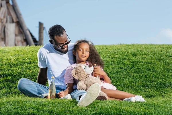 African american father and daughter sitting on hill with soda and teddy bear at amusement park — Stock Photo