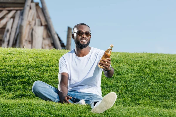 Guapo afroamericano hombre sentado en verde colina y la celebración de soda en botella de vidrio - foto de stock