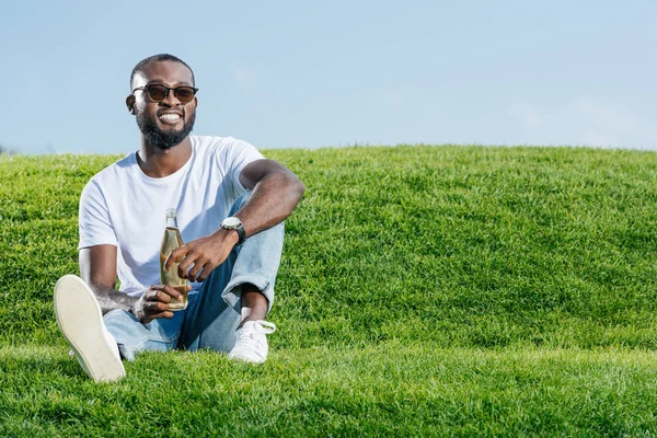 Happy handsome african american man sitting with soda in glass bottle on green hill — Stock Photo
