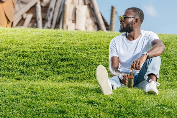 Bel homme afro-américain assis avec du soda dans une bouteille en verre sur une colline verte et regardant loin — Photo de stock