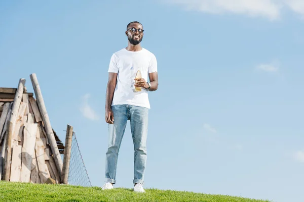 Schöner afrikanisch-amerikanischer Mann steht mit Soda in Glasflasche auf grünem Hügel — Stockfoto