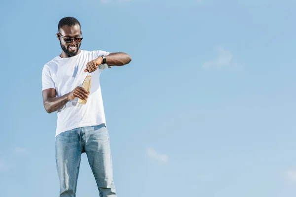Felice bello afro americano uomo aprendo soda bottiglia contro il cielo blu — Foto stock