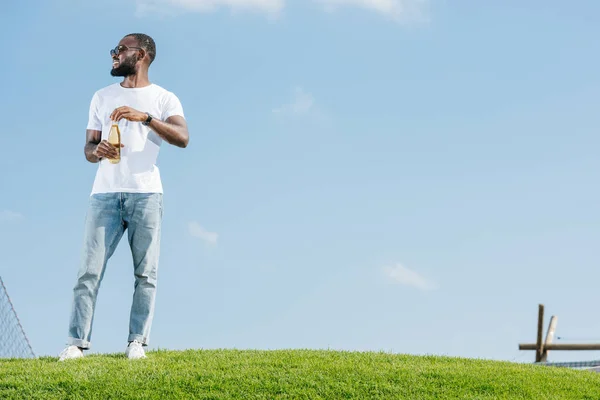 Smiling handsome african american man opening soda bottle on green hill and looking away — Stock Photo