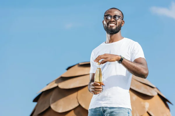 Sorridente bello africano americano uomo aprendo bottiglia di soda contro il cielo blu — Foto stock