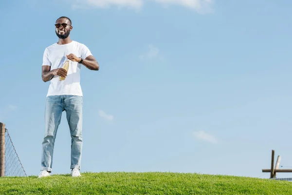 Smiling handsome african american man opening soda bottle on green hill — Stock Photo