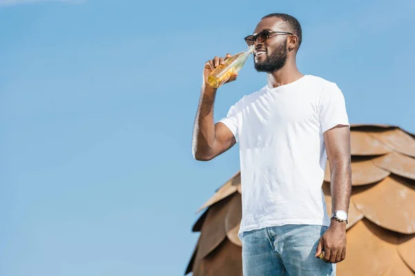 Sonriente guapo africano americano hombre bebiendo soda contra el cielo azul - foto de stock