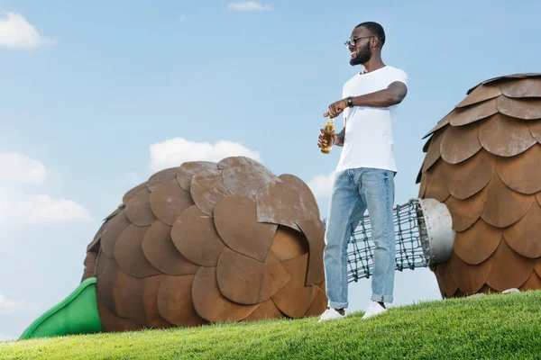 Guapo afroamericano hombre apertura botella de soda en verde colina en parque de atracciones - foto de stock