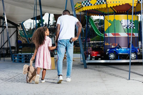 Vista trasera del policía afroamericano caminando con su hija y sosteniendo la insignia policial en el parque de atracciones - foto de stock