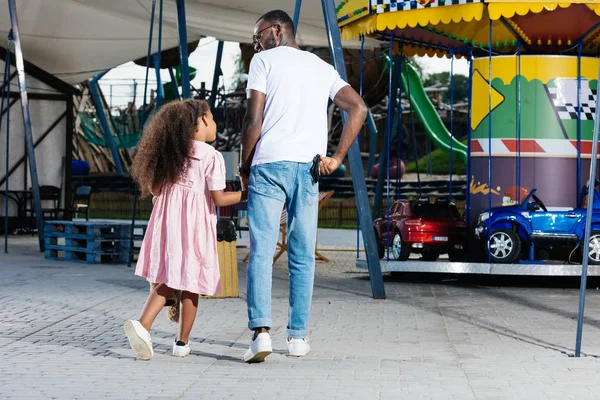 Vista trasera del policía afroamericano caminando con su hija y sosteniendo la insignia de la policía en el parque de atracciones - foto de stock