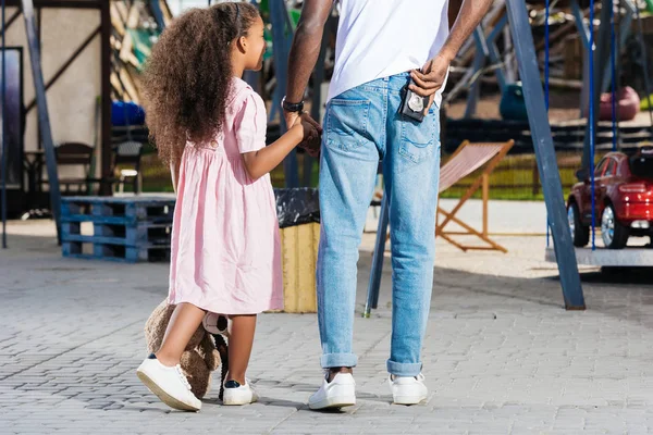 Imagen recortada del policía afroamericano caminando con su hija y sosteniendo la insignia de la policía en el parque de atracciones - foto de stock
