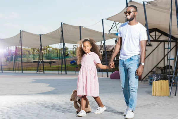 Sonrientes afroamericanos padre e hija tomados de la mano y caminando en el parque de atracciones - foto de stock