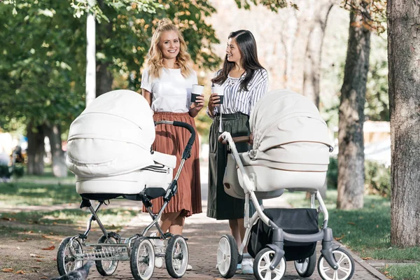 Улыбающиеся матери держат кофе, чтобы пойти и ходить с детскими колясками в парке — стоковое фото