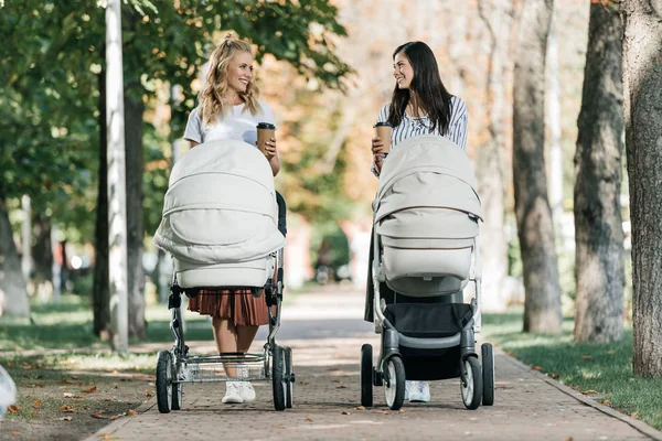 Mothers walking with baby strollers and coffee to go in park and looking at each other — Stock Photo