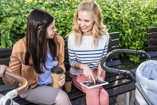 Smiling mothers sitting on bench near baby strollers with coffee to go and tablet — Stock Photo