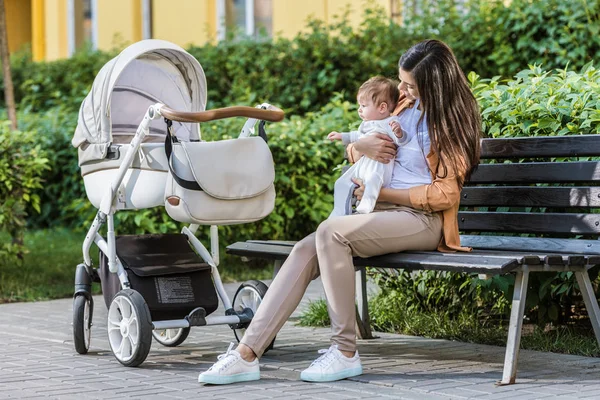 Mãe sentada com o bebê no banco perto do carrinho no parque — Fotografia de Stock
