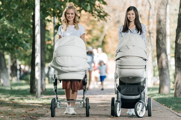 Sonrientes madres caminando con cochecitos de bebé en el parque y mirando a la cámara - foto de stock