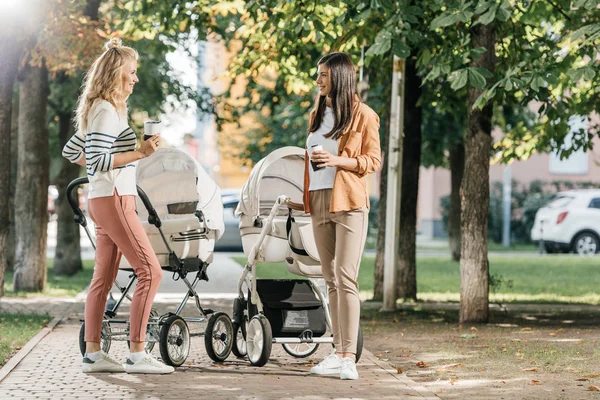 Mothers standing with coffee to go near baby strollers in park — Stock Photo
