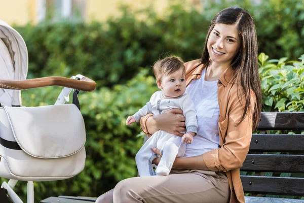 Smiling mother holding daughter near stroller in park and looking at camera — Stock Photo